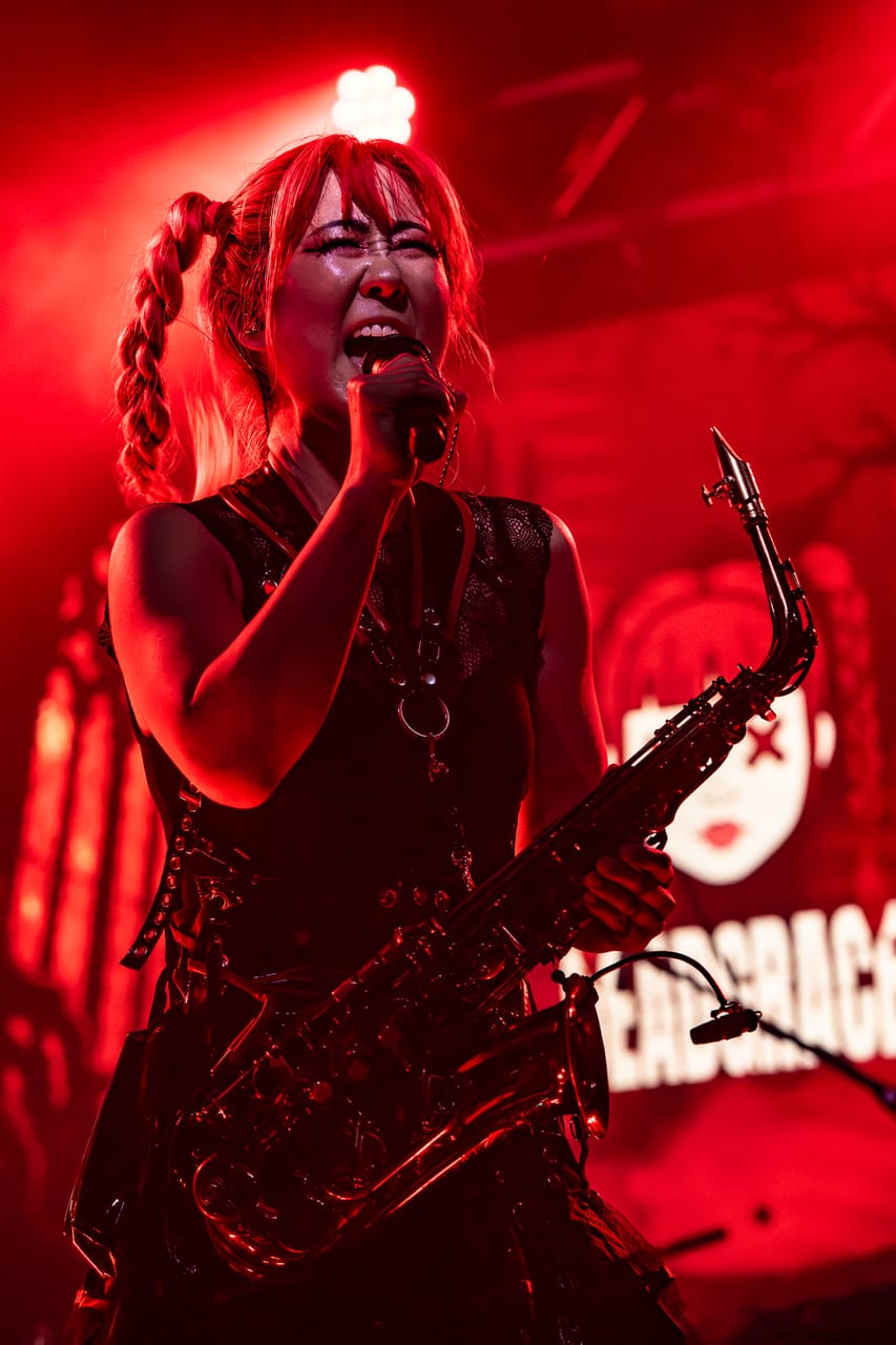Grace Kelly sings into a microphone with a saxophone in her other hand, bathed in red light in front of the DEADGRACE logo in Albuquerque