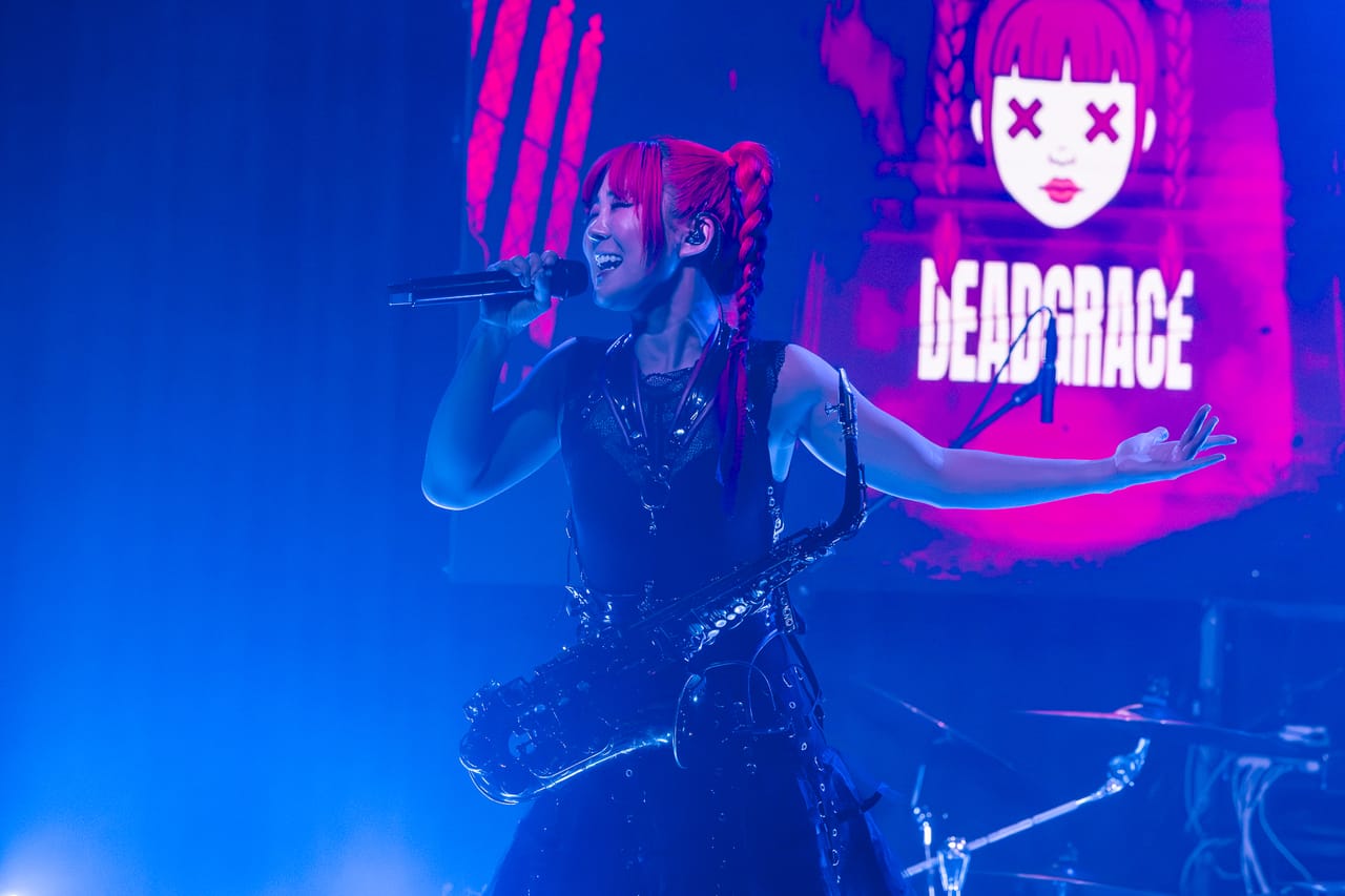 Grace Kelly sings with her arm outstretched and a saxophone on its strap in blue light, the DEADGRACE logo on the screen behind her in Albuquerque