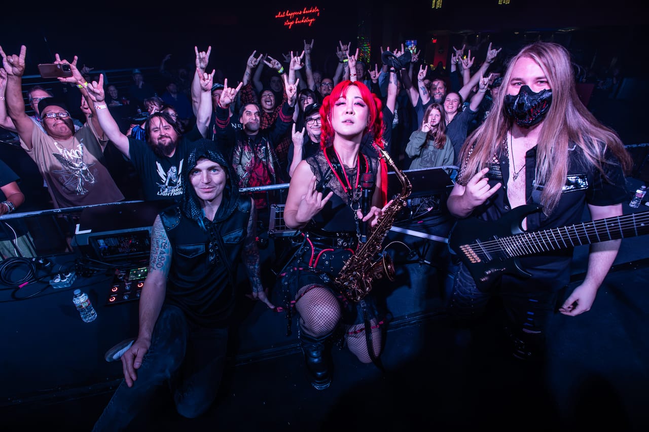 Kyle Brian, Grace Kelly with her saxophone, and Sam Fairless of DEADGRACE kneel at the front of the stage in Albuquerque, the crowd behind them throwing horns