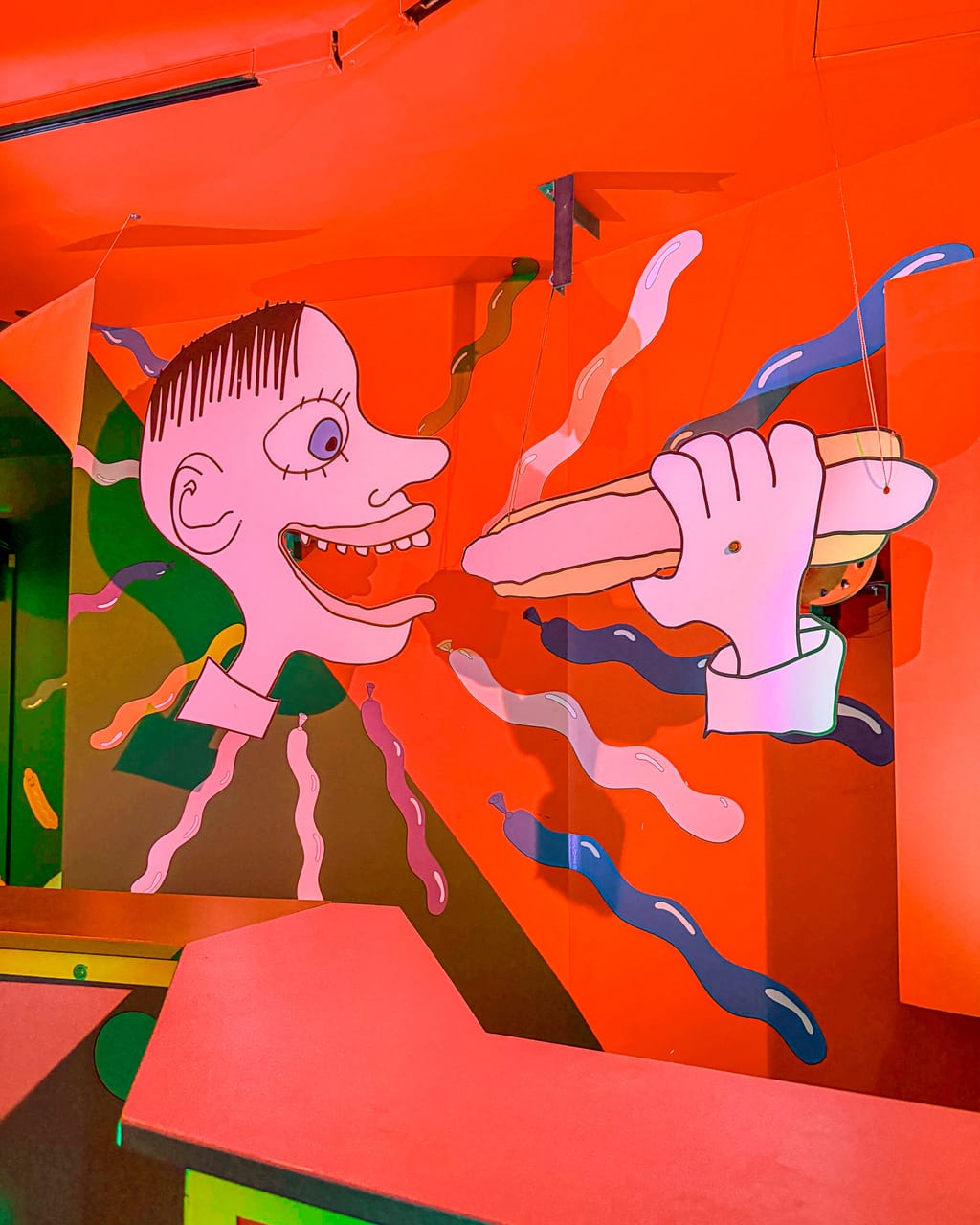 A bright red room at Meow Wolf in Denver painted with a cartoon face reaching for a hot dog, wavy ribbons around it