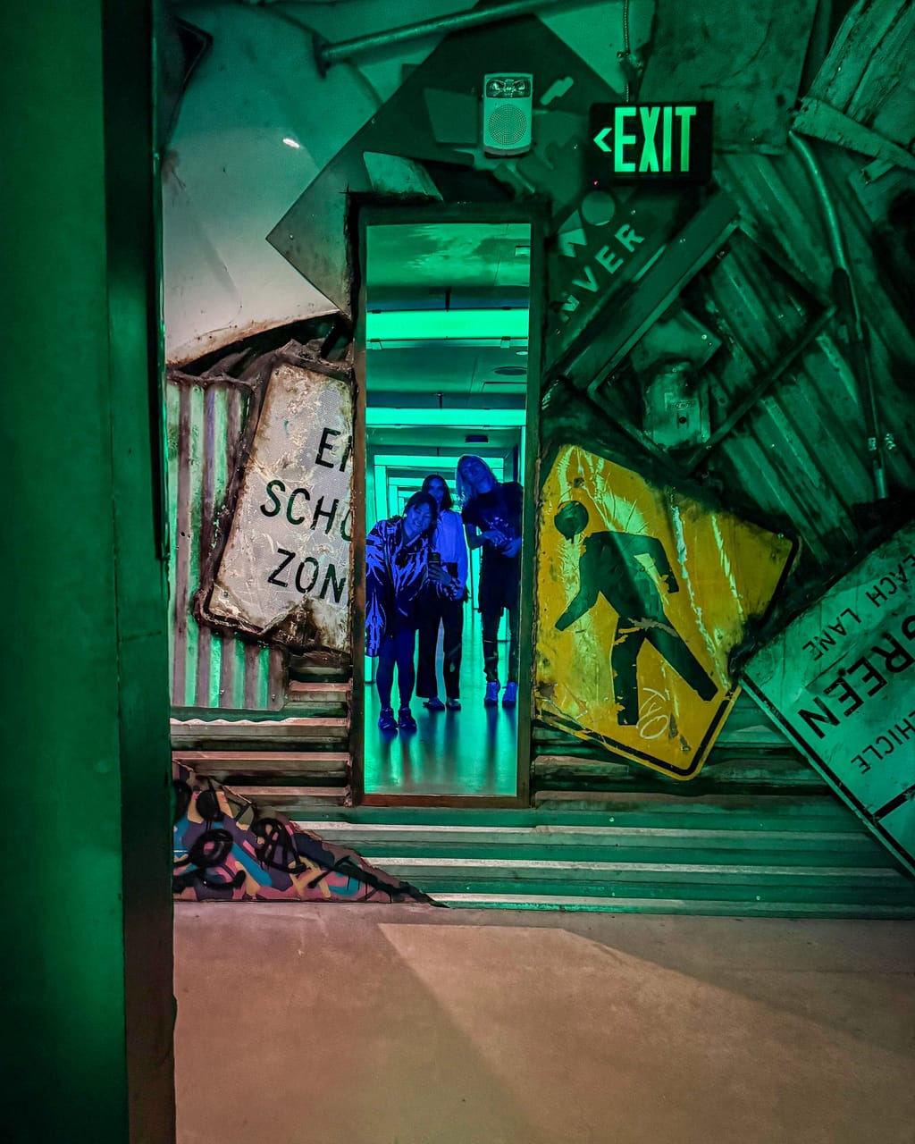 A glowing green exit doorway framed by bent road signs inside Meow Wolf in Denver, people walking through into the next room