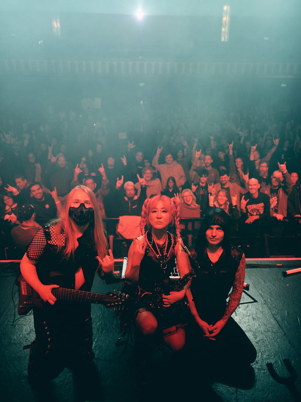 Sam Fairless, Grace Kelly and Kyle Brian of DEADGRACE kneel at the front of the stage at the Oriental Theater, the Denver crowd throwing horns behind them