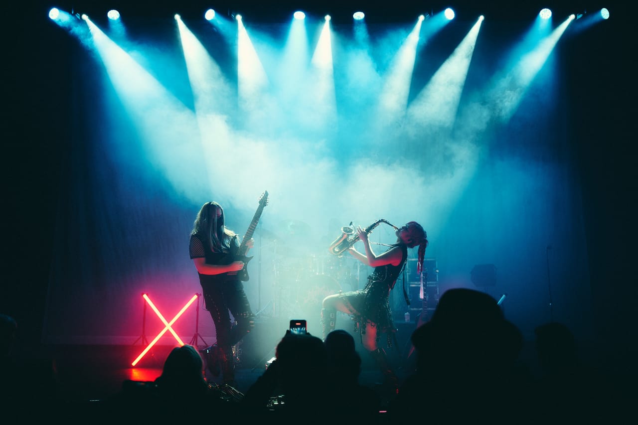 Wide shot of DEADGRACE on stage at the Oriental Theater in Denver: Sam Fairless on guitar and Grace Kelly on saxophone under a fan of blue spotlights, a neon X glowing red, the crowd in silhouette
