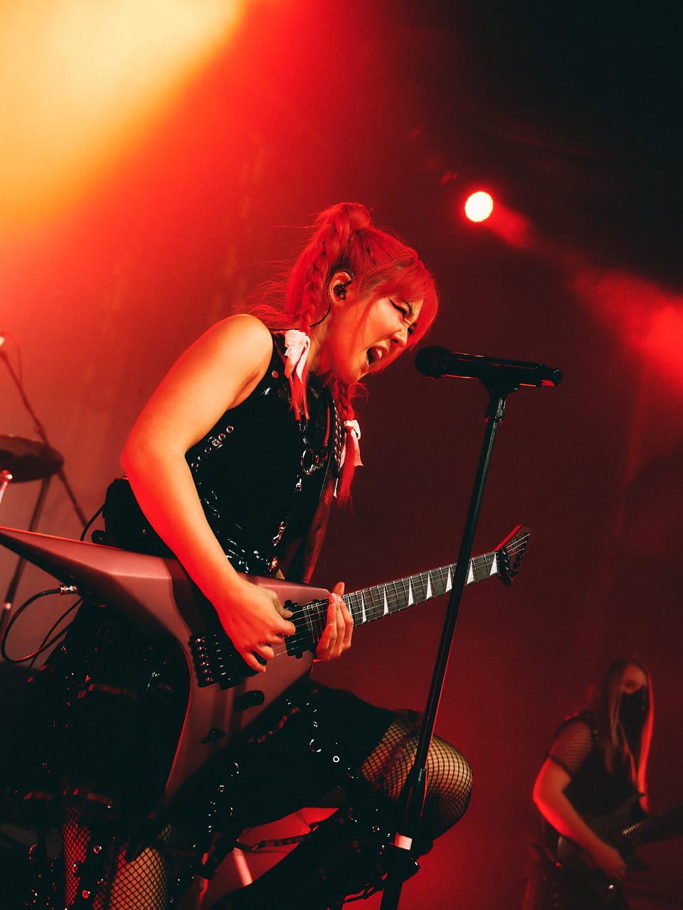 Grace Kelly screams into a microphone stand while playing a flying-V guitar under red light at the Oriental Theater in Denver, Sam Fairless behind her