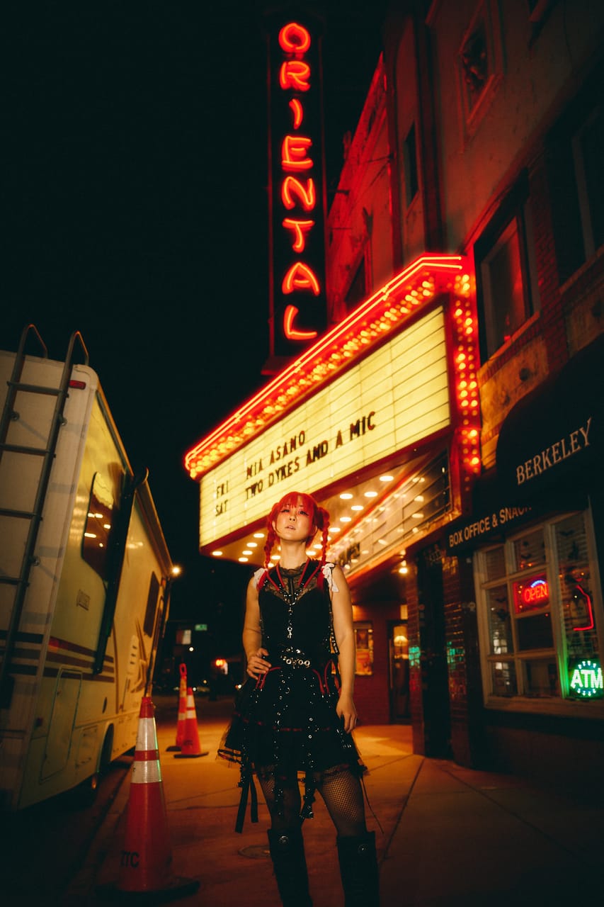 Grace Kelly of DEADGRACE stands on the sidewalk under the lit Oriental Theater marquee and vertical neon sign in Denver at night, an RV parked at the curb