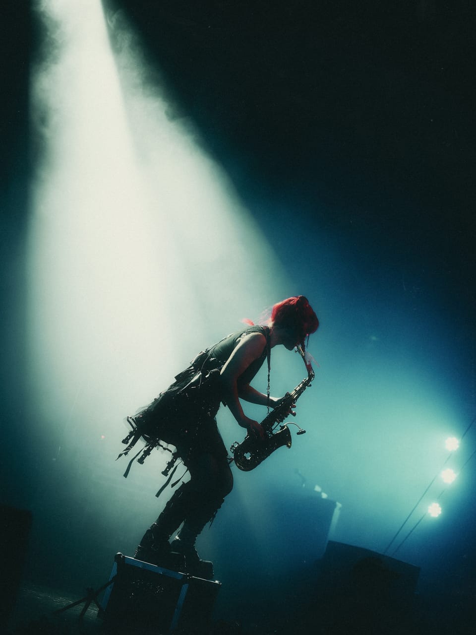 Grace Kelly in silhouette plays saxophone with one boot up on a road case, a beam of light cutting through haze at the Oriental Theater, Denver
