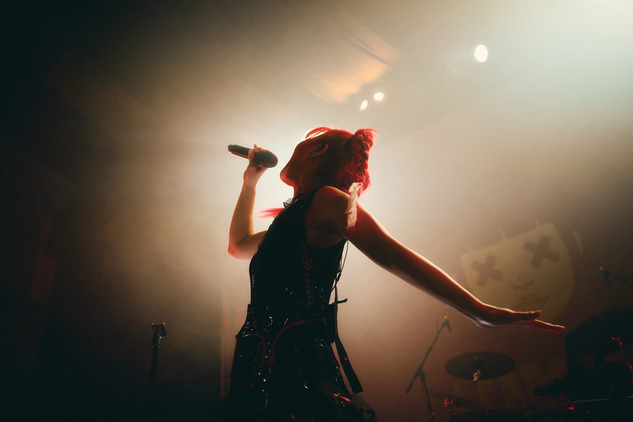 Grace Kelly sings into a handheld microphone with her other arm outstretched, backlit through haze on stage at the Oriental Theater, Denver