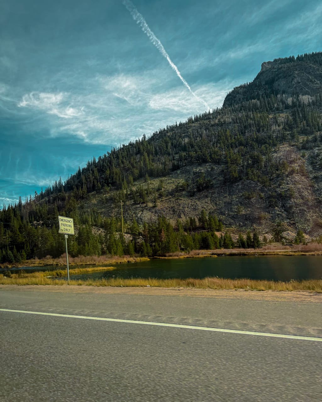 A mountain highway in Colorado on the drive to Grand Junction: pine-covered slopes, still water by the road and a contrail across a blue sky