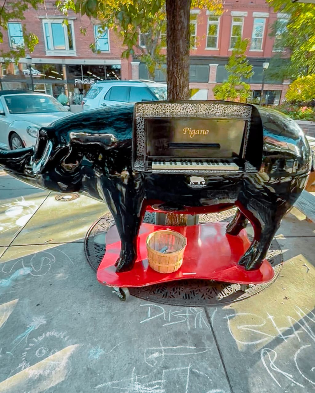 A black pig-shaped street piano labeled Pigano on a sidewalk in downtown Grand Junction, chalk drawings on the pavement around it