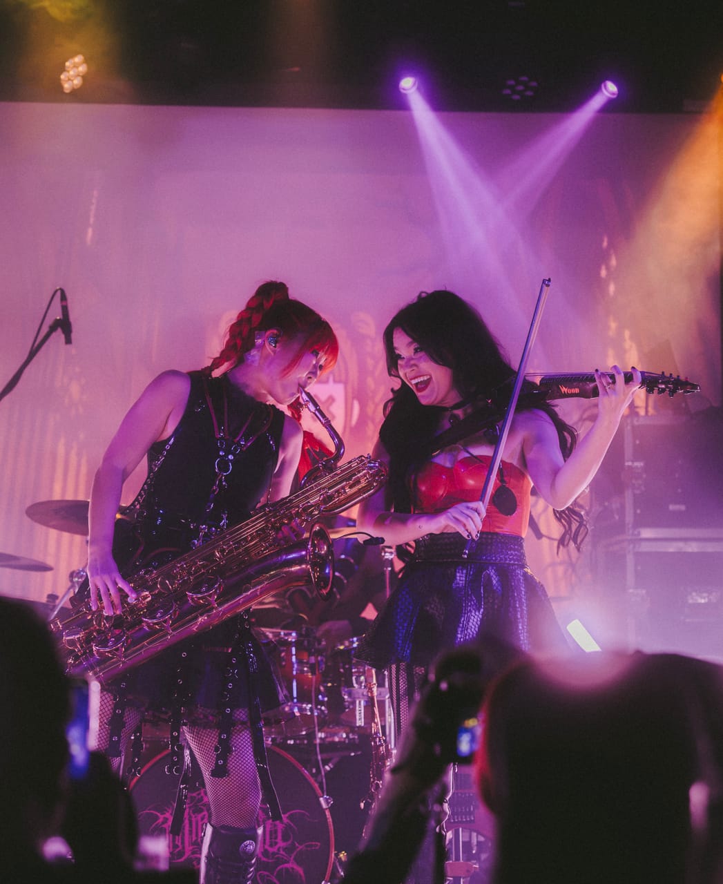 Grace Kelly on baritone saxophone and Mia Asano on electric violin laugh together on stage under pink and purple light at the Star Theater, phones raised in the crowd