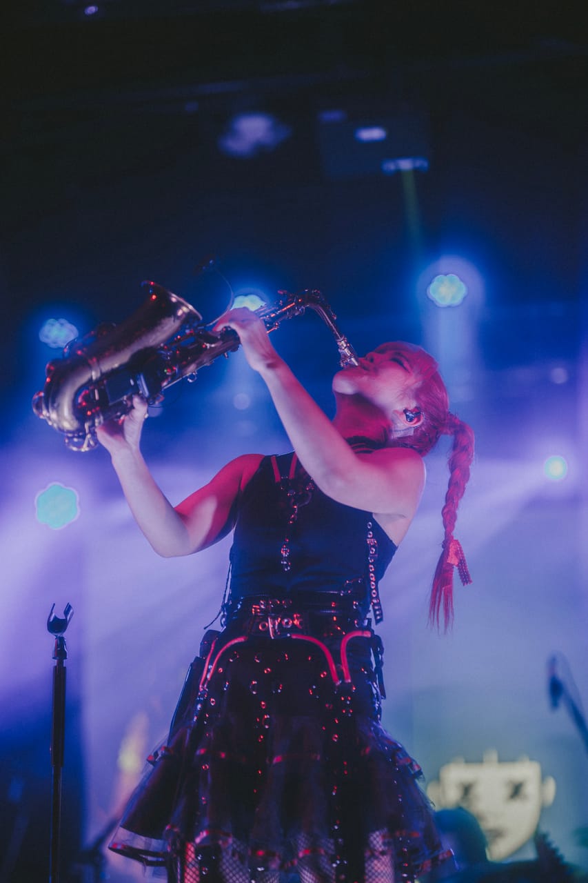 Low-angle photo of Grace Kelly playing saxophone with the bell raised under blue stage light at the Star Theater, the DEADGRACE logo on the screen behind her