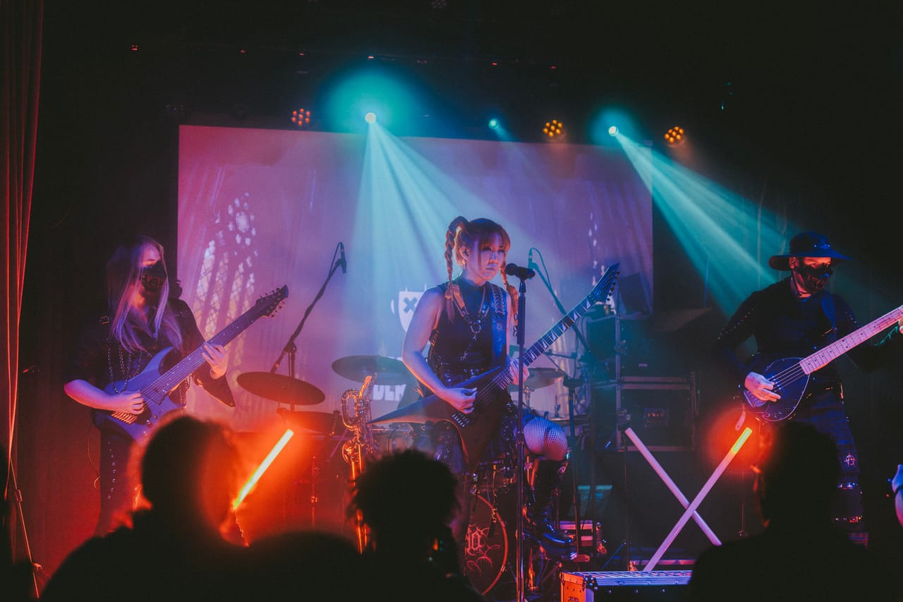 DEADGRACE on stage at the Star Theater: Grace Kelly sings and plays guitar seated in the middle, masked guitarist Sam Fairless on the left and bassist Hampus Isaksson on the right, under teal and pink lights
