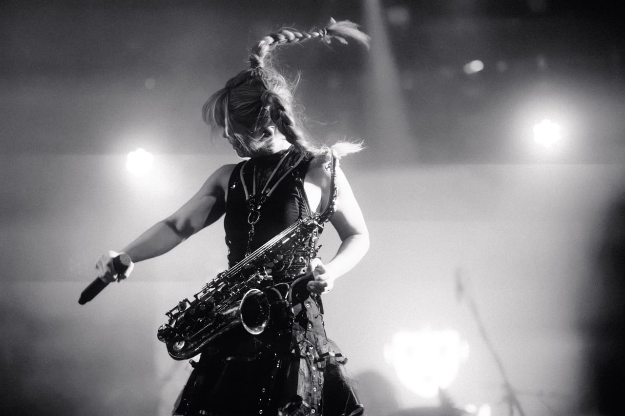 Black-and-white photo of Grace Kelly mid-headbang at the Star Theater, a saxophone on its strap and a microphone in hand, one braid flying above her head