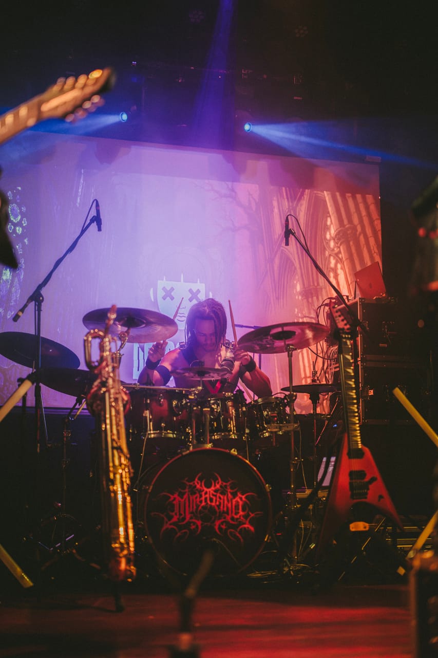 Dane White plays drums at the Star Theater behind a kit with Mia Asano's logo on the kick drum, a baritone saxophone and a red flying-V guitar on stands in front