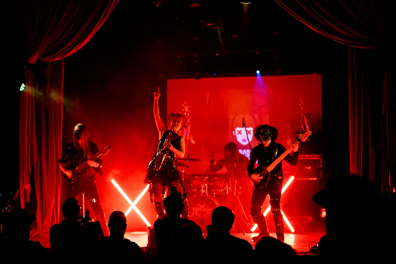 Wide photo of the Star Theater stage bathed in red light and framed by red velvet curtains: Sam Fairless on guitar, Grace Kelly with a saxophone and one arm raised, Dane White on drums, and Hampus Isaksson on bass, the crowd in silhouette