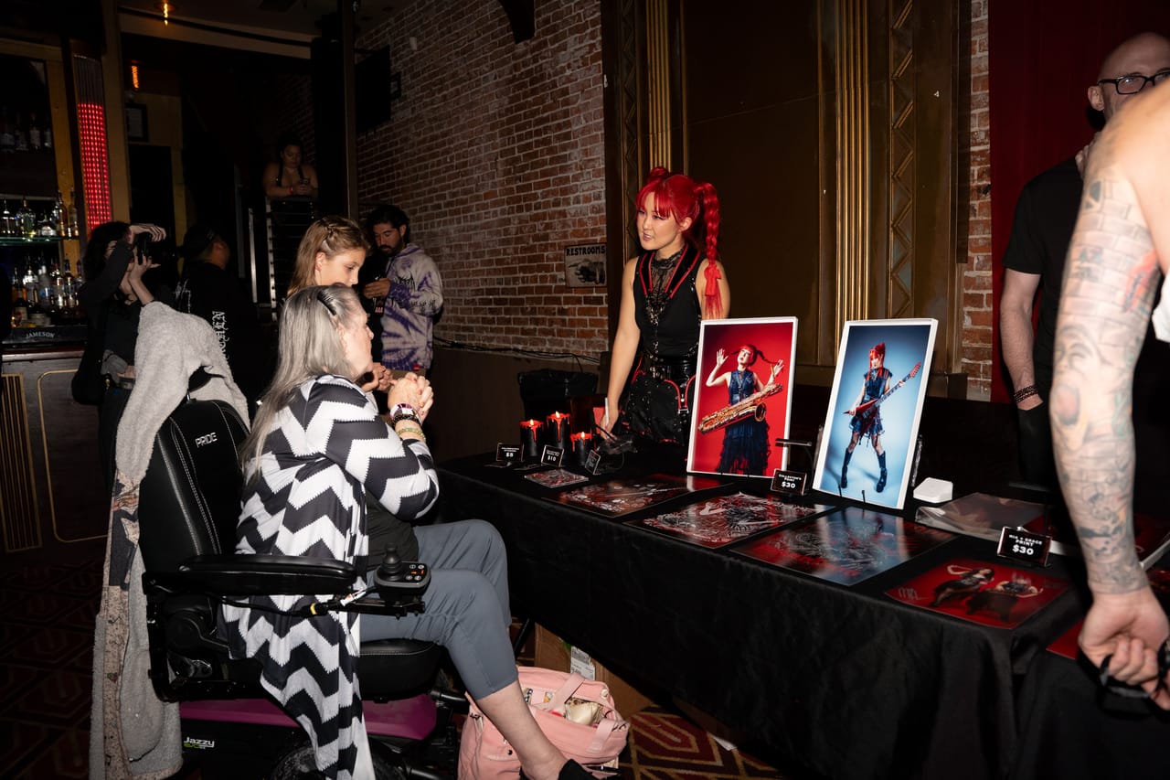 Grace Kelly behind the DEADGRACE merch table after the set, talking with fans, framed prints and red candles on the table and a brick wall with gold art-deco trim behind