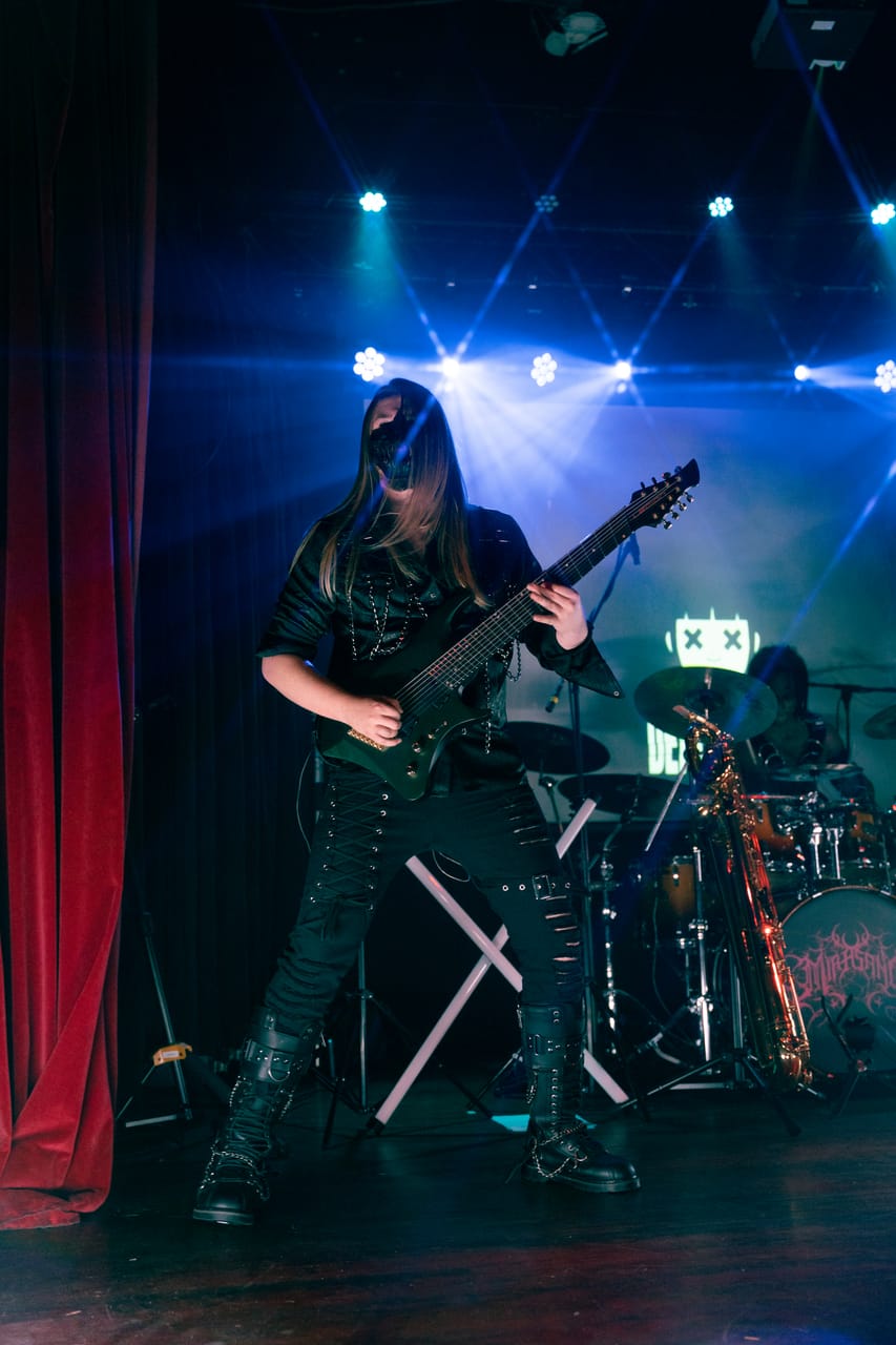 DEADGRACE guitarist Sam Fairless, long blond hair and a black mask, plays an eight-string guitar beside the red velvet curtain at the Star Theater, a baritone saxophone and the drum kit behind