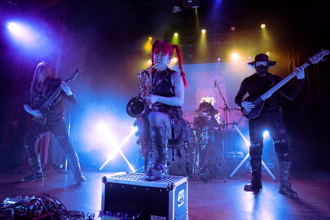 Grace Kelly sits on a road case at the front of the Star Theater stage holding a saxophone, between Sam Fairless on guitar and Hampus Isaksson on bass, in blue and gold light and haze
