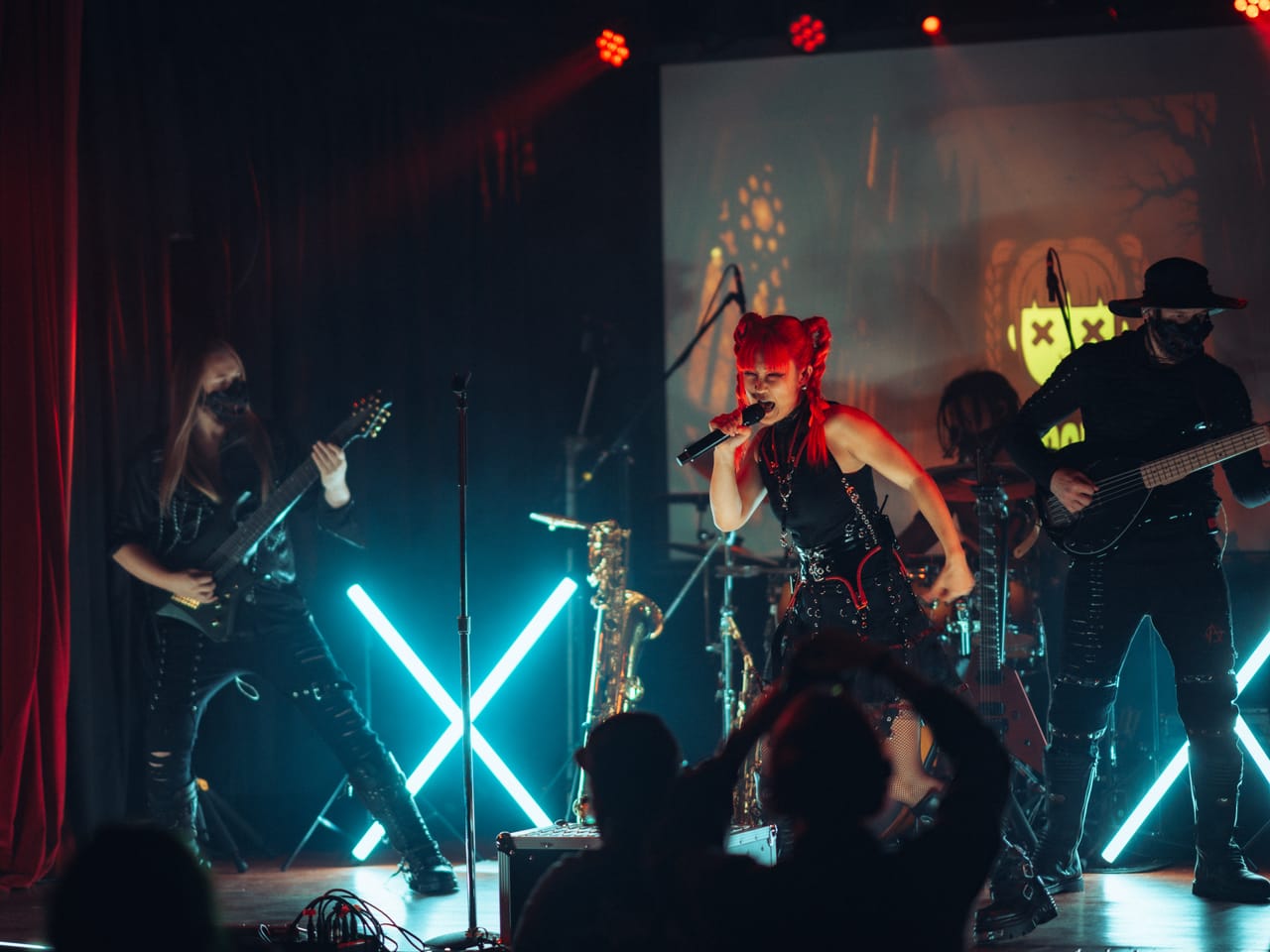 DEADGRACE live at the Star Theater: Grace Kelly sings with a fist clenched between two teal neon X lights, Sam Fairless on guitar to the left and Hampus Isaksson in a black hat on bass to the right, the crowd in the foreground