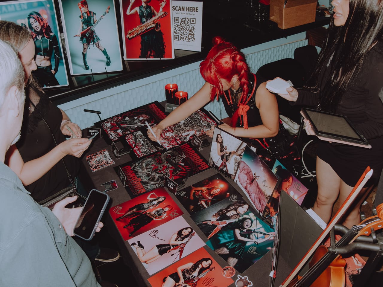 Grace Kelly signing prints at the merch table at Harlow's while fans look over posters and photo prints, a merch seller holding a tablet beside her