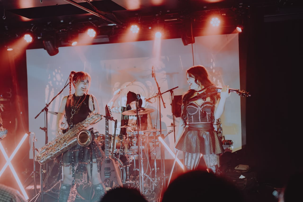 Grace Kelly with her saxophone and violinist Mia Asano laughing together on stage at Harlow's in Sacramento, drummer Kyle Brian in a hood behind the kit and a projection screen behind them