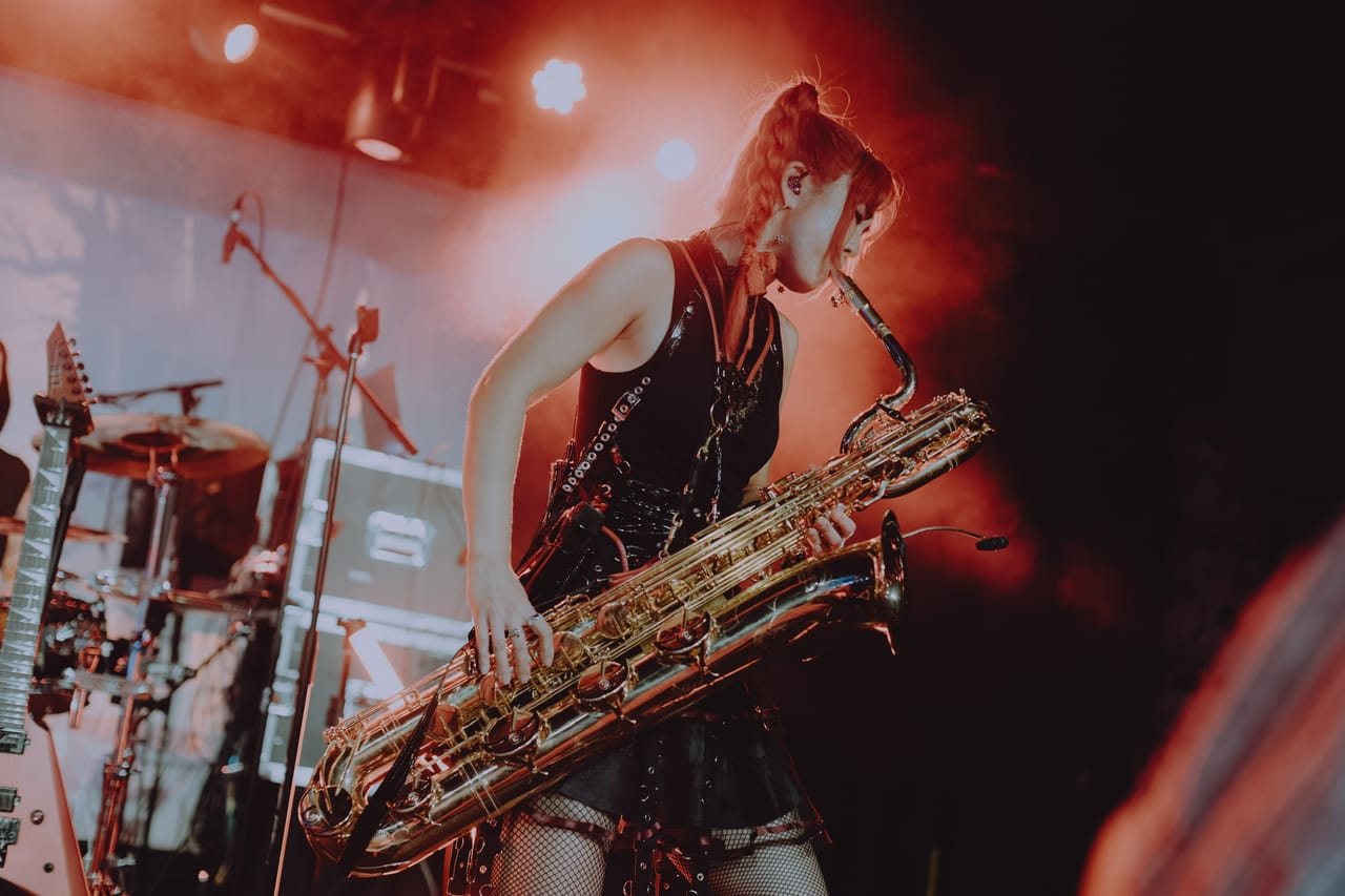 Grace Kelly plays baritone saxophone under red stage light at Harlow's in Sacramento, a flying-V guitar on a stand beside the drum kit