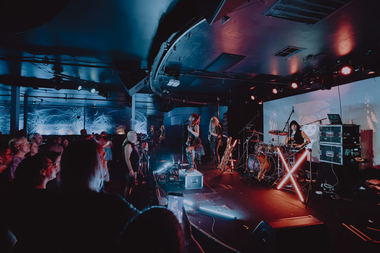 Wide shot of DEADGRACE live at Harlow's in Sacramento: Grace Kelly at the front of the stage, masked guitarist Sam Fairless beside her, drummer Kyle Brian behind the kit and a neon X light, with the crowd watching and filming from the floor