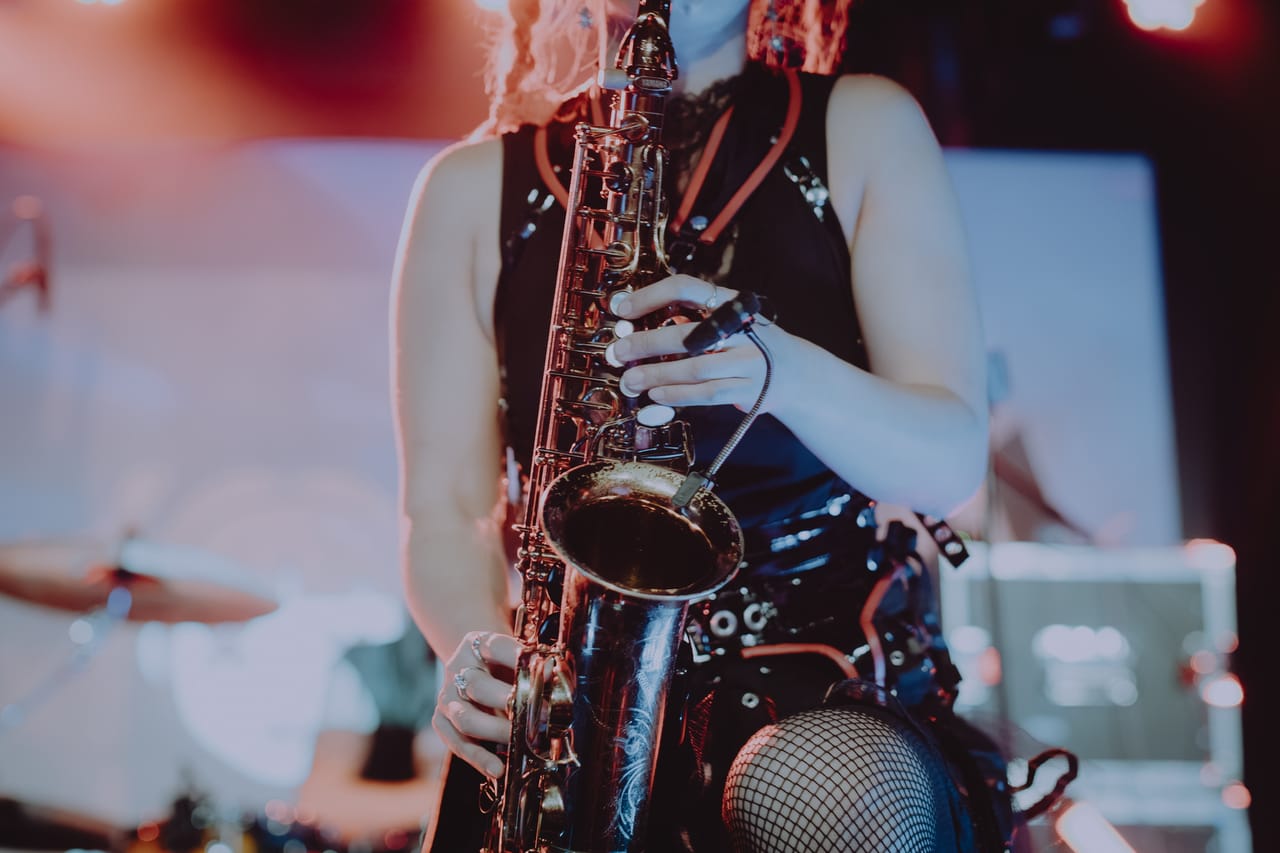 Close-up of Grace Kelly's hands on a saxophone with a clip-on microphone on the bell, studded belts and fishnets behind it, on stage at Harlow's
