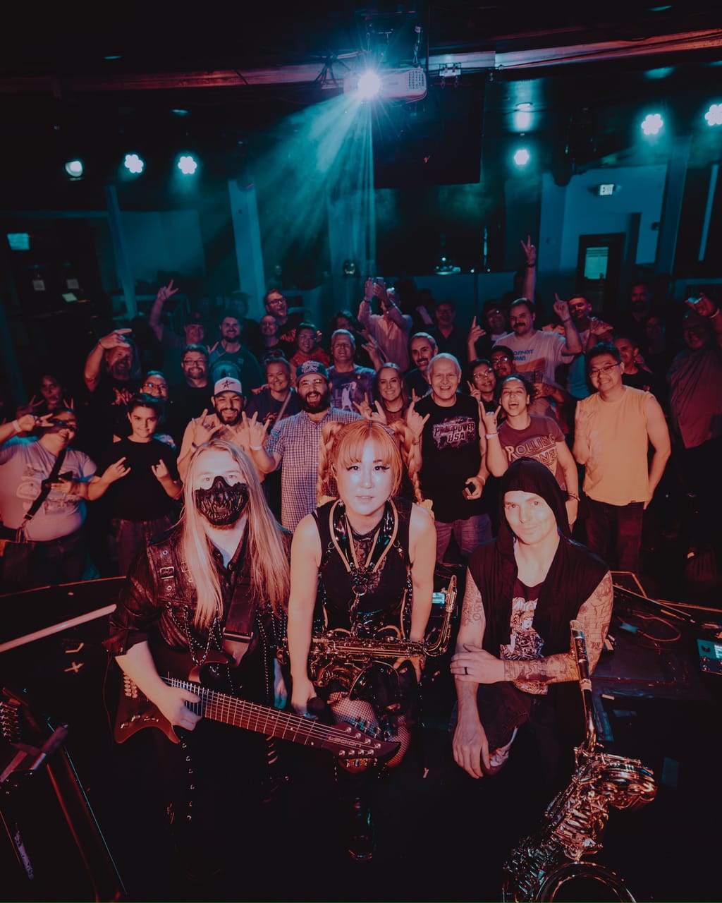 DEADGRACE posing on stage at Harlow's in Sacramento — masked guitarist Sam Fairless with an 8-string guitar, Grace Kelly with her saxophone, and drummer Kyle Brian in a hood — with the crowd behind them throwing horns