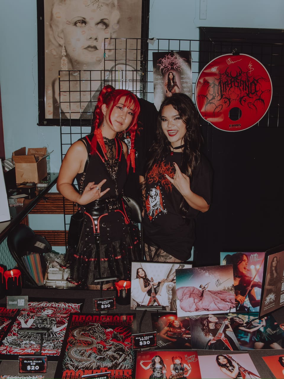 Grace Kelly and Mia Asano at the merch table at Harlow's, Grace throwing horns, under a vintage glamour portrait and a large Mia Asano CD, with DEADGRACE and Mia x Grace prints on the table