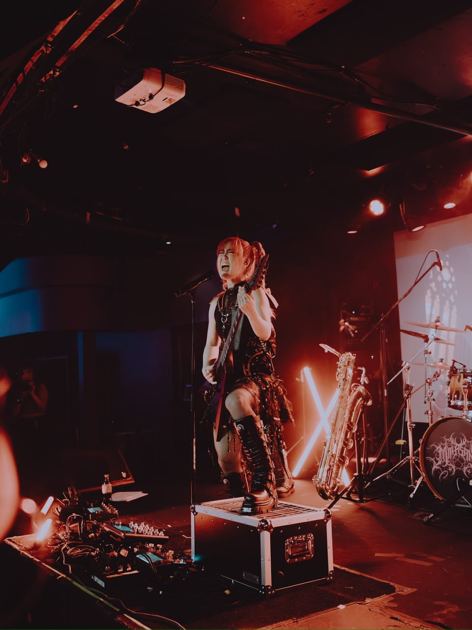 Grace Kelly of DEADGRACE sings and plays guitar standing on a road case at Harlow's, Sacramento, with a baritone saxophone, a neon X light, and the drum kit behind her