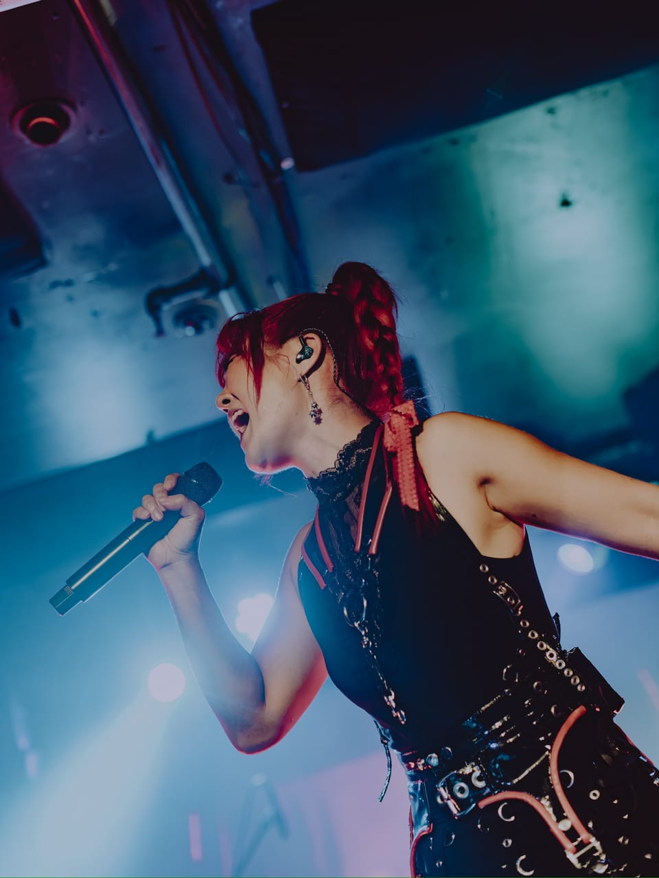Low-angle close-up of Grace Kelly of DEADGRACE singing into a handheld microphone under teal light at Harlow's, Sacramento, red braid and in-ear monitor visible