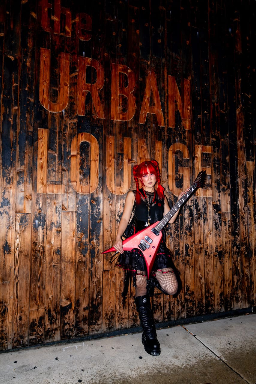 Grace Kelly of DEADGRACE stands on one leg holding a red flying-V guitar against the weathered wooden wall painted with the Urban Lounge sign in Salt Lake City