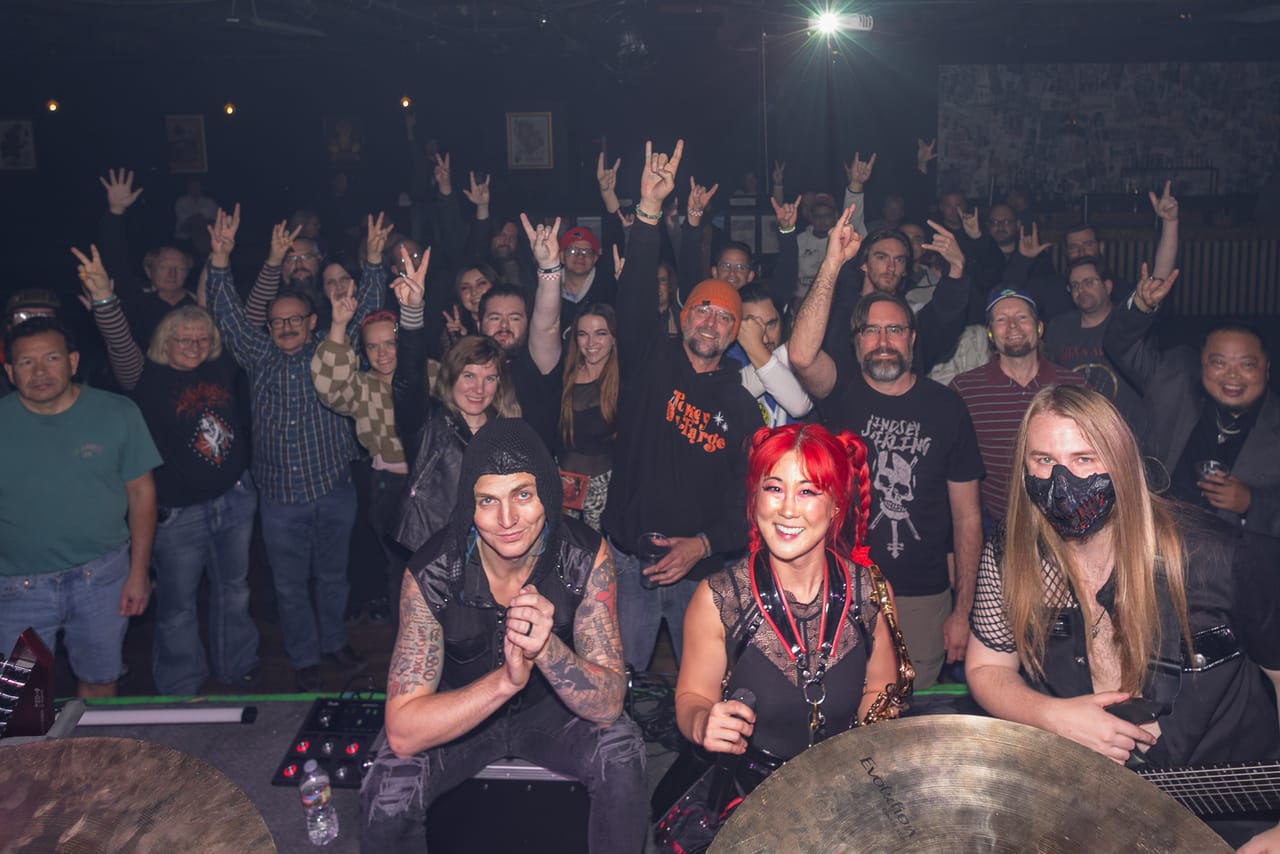 Kyle Brian, Grace Kelly and Sam Fairless of DEADGRACE pose at the front of the stage at the Urban Lounge in Salt Lake City, the crowd behind them throwing horns