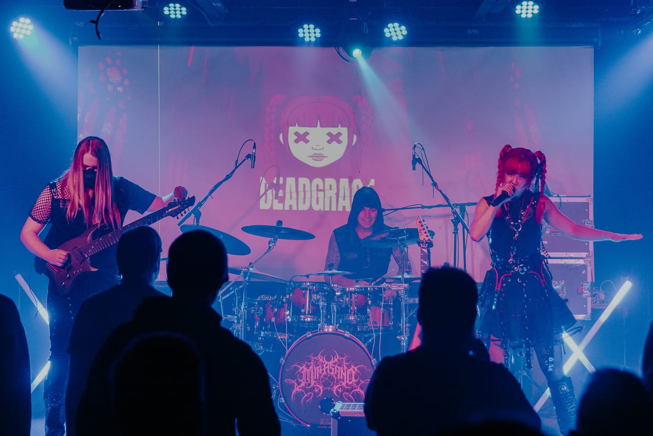 DEADGRACE on stage at the Urban Lounge in Salt Lake City, seen over the heads of the crowd: guitarist Sam Fairless, drummer Kyle Brian and Grace Kelly singing with one arm out, the DEADGRACE logo on the screen behind them