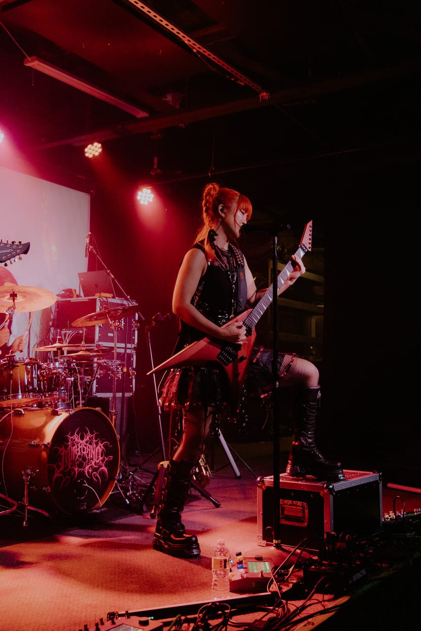 Grace Kelly plays a guitar with one boot up on a road case under red light at the Urban Lounge in Salt Lake City, the drum kit behind her
