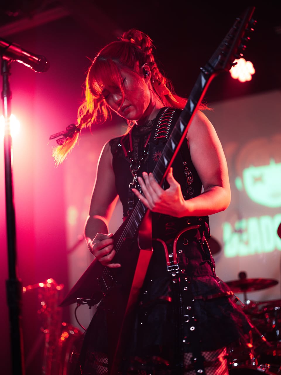 Grace Kelly looks down at the neck of her flying-V guitar under magenta light at the Urban Lounge, the DEADGRACE logo glowing on the screen behind her