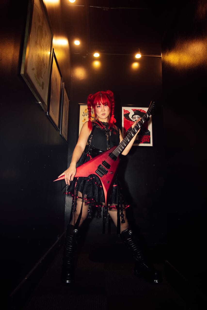 Grace Kelly poses with a red flying-V guitar in a narrow backstage hallway lined with framed gig posters at the Urban Lounge, Salt Lake City