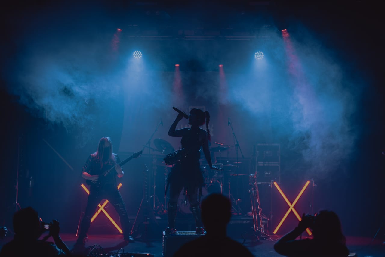 Silhouette of Grace Kelly singing into a handheld mic from a road case, guitarist Sam Fairless and the drum kit behind her, framed by two orange neon X lights and blue haze while fans film from the floor