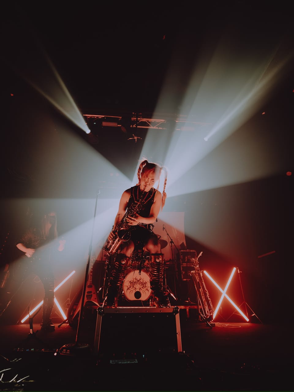 Grace Kelly plays saxophone from a road case in front of the drum kit as white light beams fan out overhead, with a baritone saxophone on a stand and neon X lights at either side