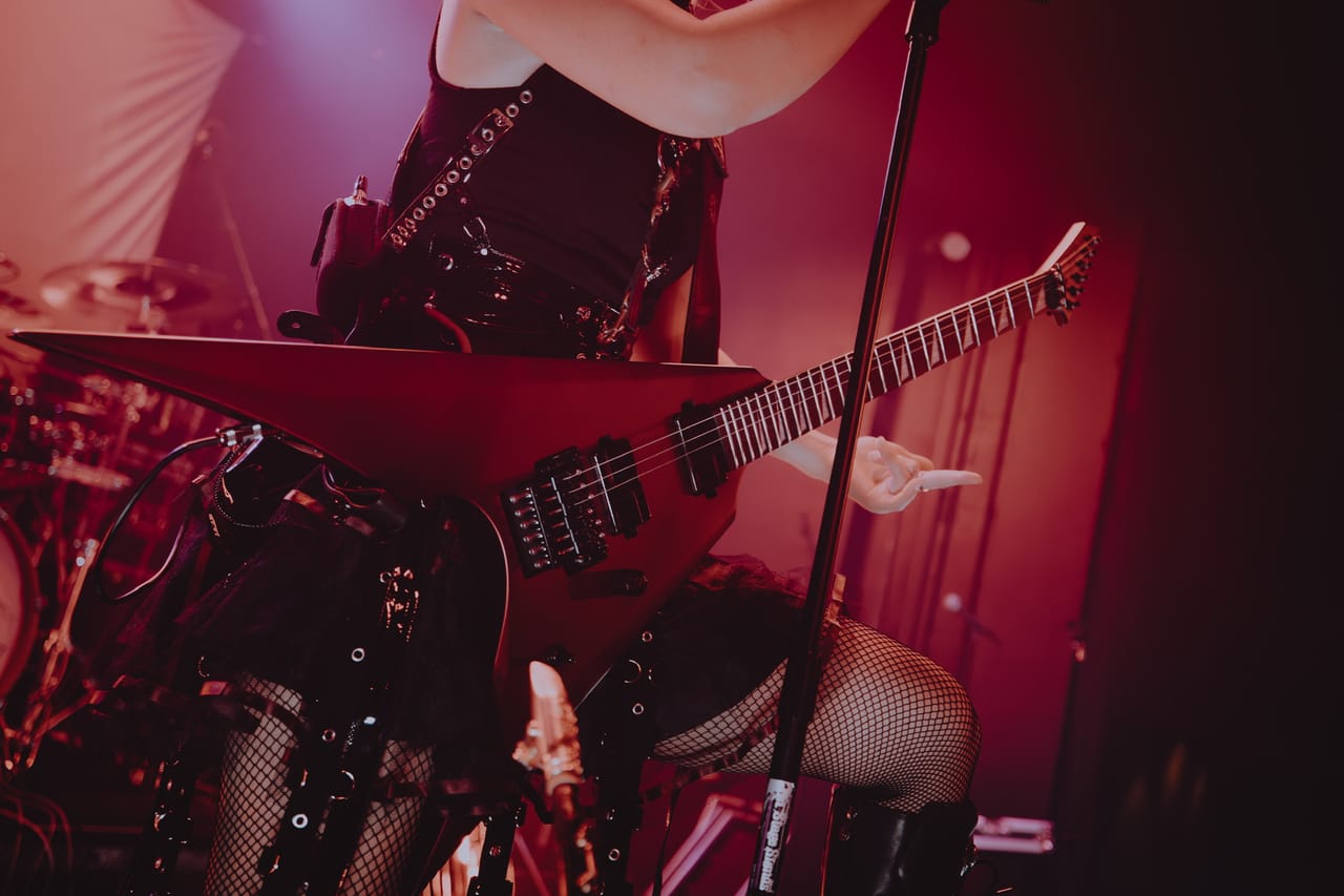 Close-up of Grace Kelly's red flying-V guitar, fishnets, and studded belts beside a mic stand, with the drum kit glowing pink behind