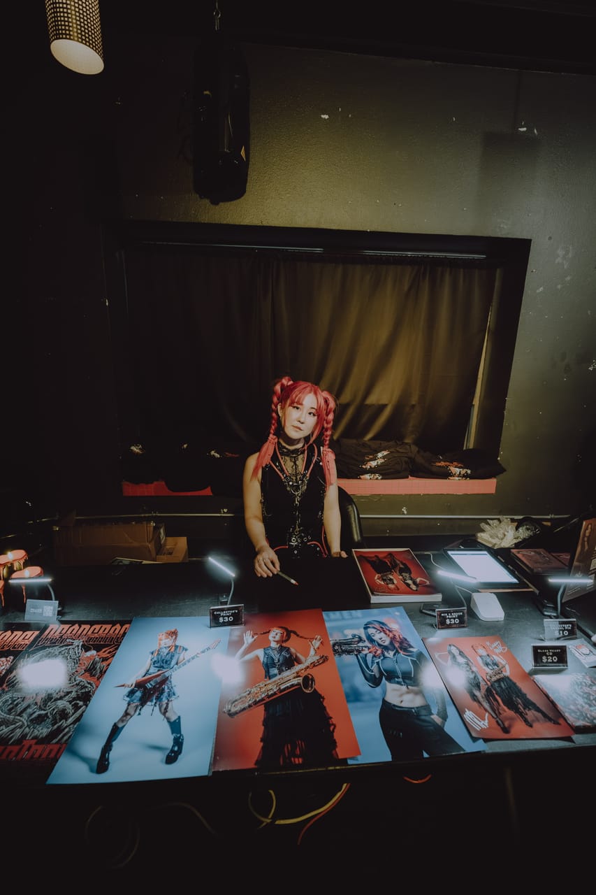 Grace Kelly at the DEADGRACE merch table at The Ritz, behind a row of $30 posters showing her with a flying-V guitar and a baritone saxophone