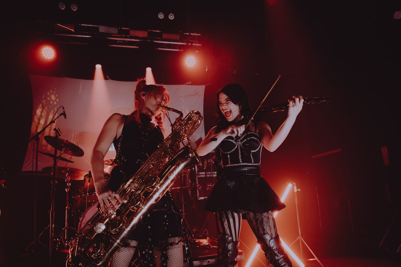 Grace Kelly plays baritone saxophone next to violinist Mia Asano, who shouts with her bow raised, under red lights on stage at The Ritz