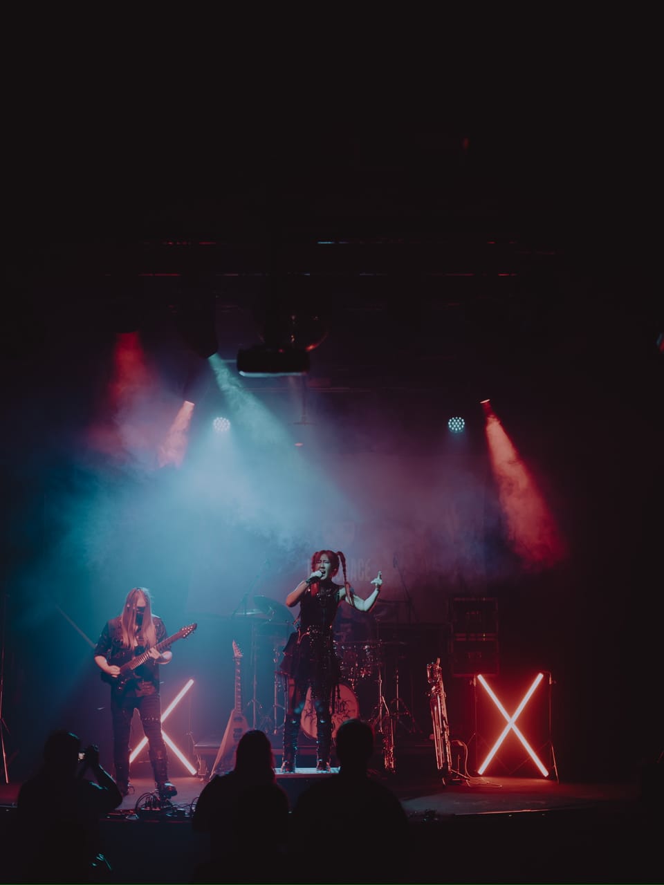 Wide shot of DEADGRACE on stage at The Ritz in San Jose: Grace Kelly sings with one arm raised, masked guitarist Sam Fairless plays stage left, a flying-V guitar and a saxophone wait on stands, and the crowd is silhouetted in front
