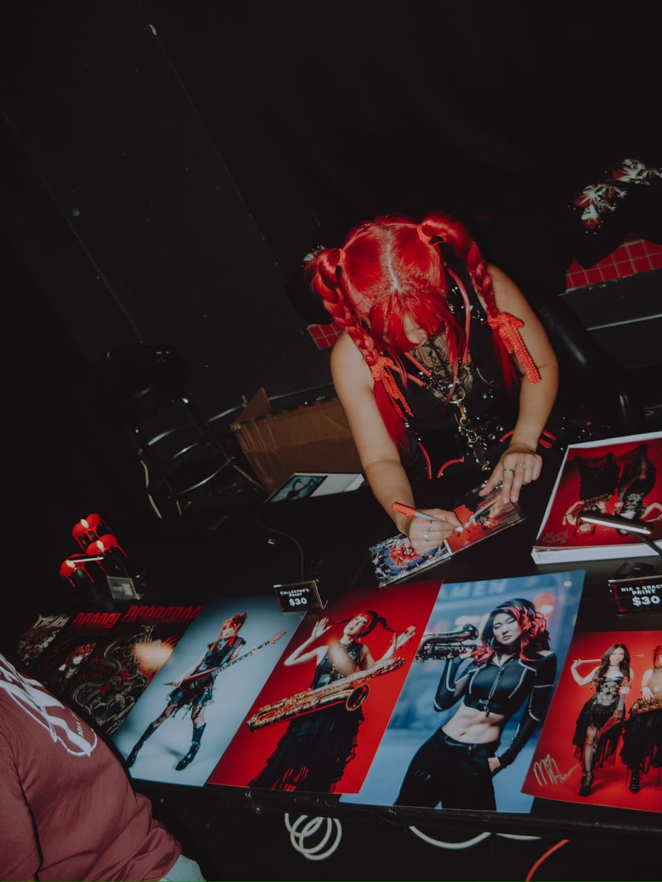 Grace Kelly signing prints at the merch table, with posters of her on flying-V guitar and baritone saxophone and a Mia x Grace print laid out in front