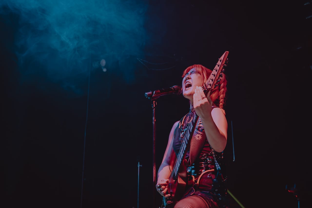 Grace Kelly of DEADGRACE, red braids and glasses, screams into the microphone while playing guitar under blue stage haze at The Ritz, San Jose