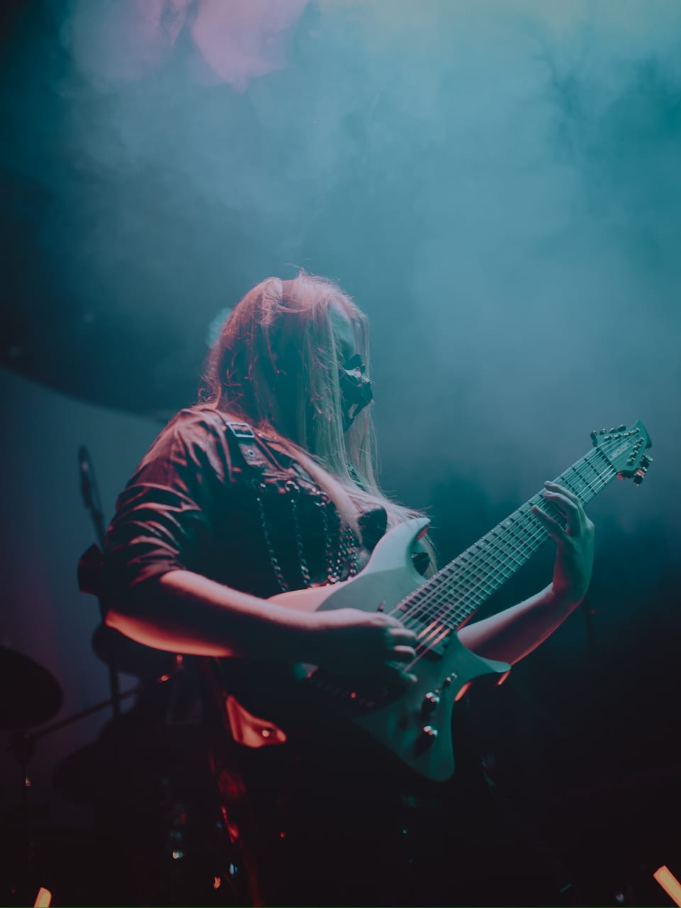 DEADGRACE guitarist Sam Fairless, long blond hair falling over a black face mask, plays an 8-string guitar in teal stage smoke at The Ritz