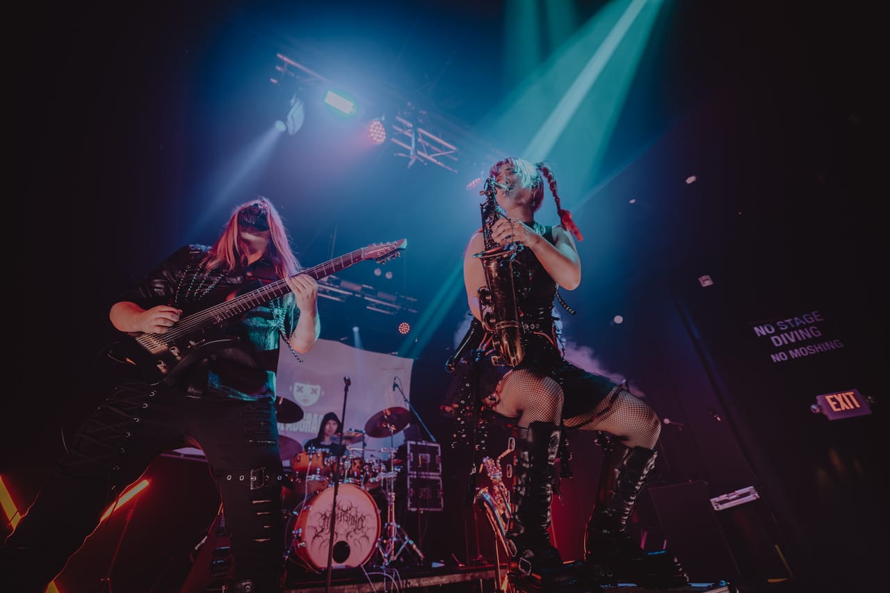 Low-angle shot of DEADGRACE guitarist Sam Fairless and Grace Kelly lifting her saxophone, drummer Kyle Brian behind them, with a NO STAGE DIVING / NO MOSHING sign glowing on the wall of The Ritz