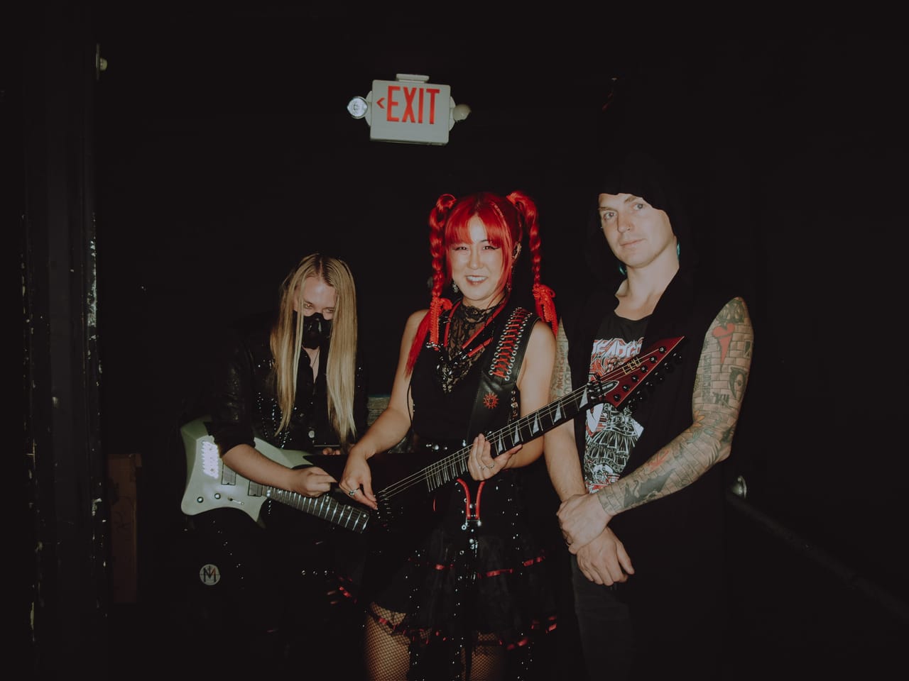 DEADGRACE under a red EXIT sign backstage at The Ritz: masked guitarist Sam Fairless holding a white guitar, Grace Kelly with her red flying-V guitar, and drummer Kyle Brian in a beanie, tattooed arms crossed