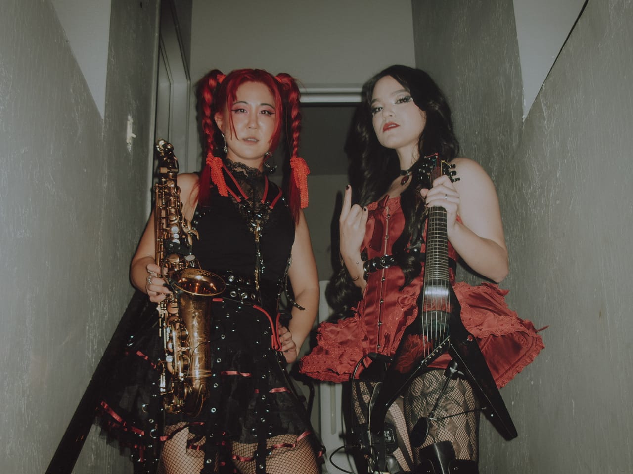 Grace Kelly holding a gold alto saxophone in a narrow stairwell at The Ritz beside violinist Mia Asano, in a red corset, throwing horns and holding a black V-shaped instrument